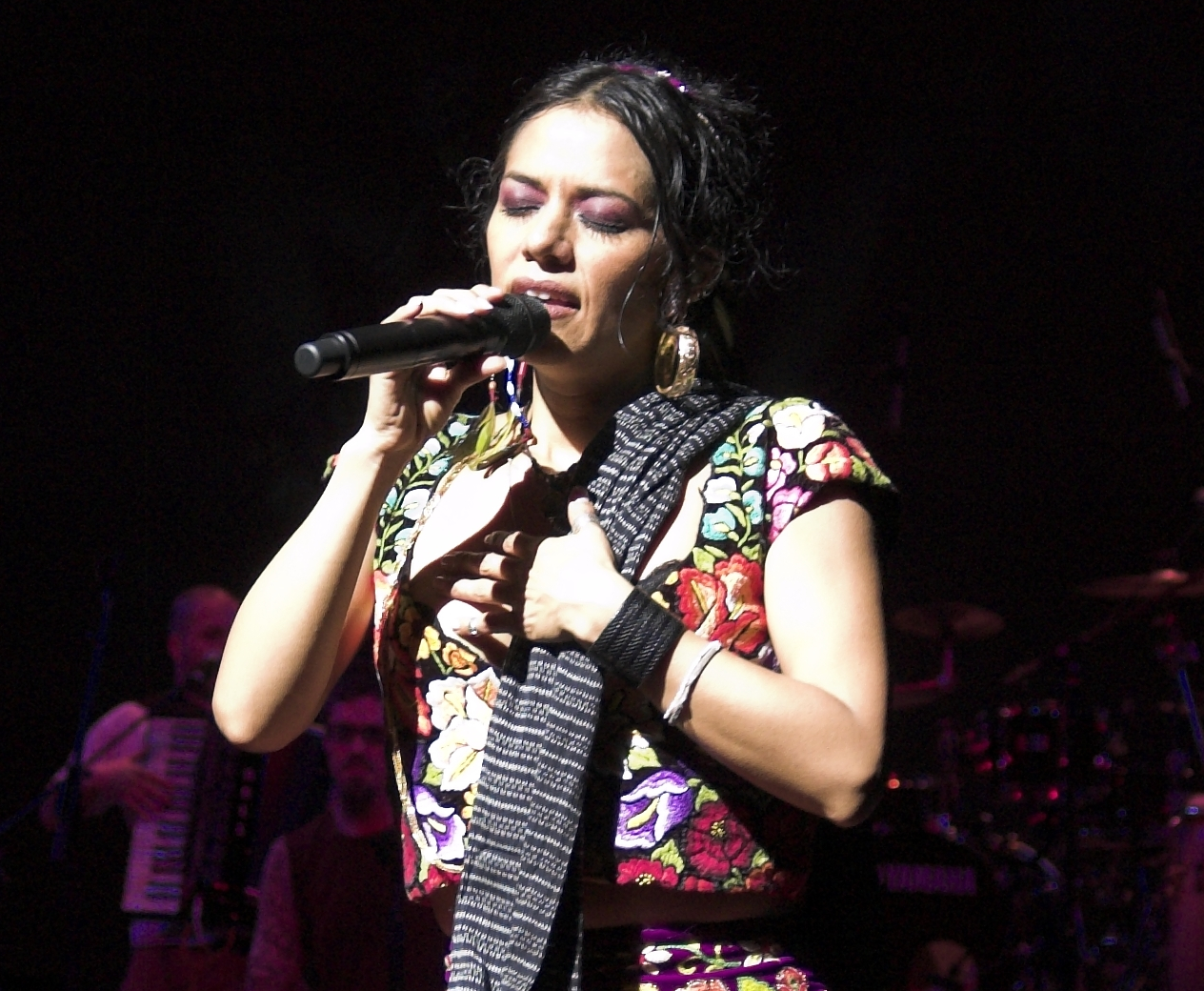Lila Downs in a black dress with colorful flowers and a long gray scarf, holding a microphone