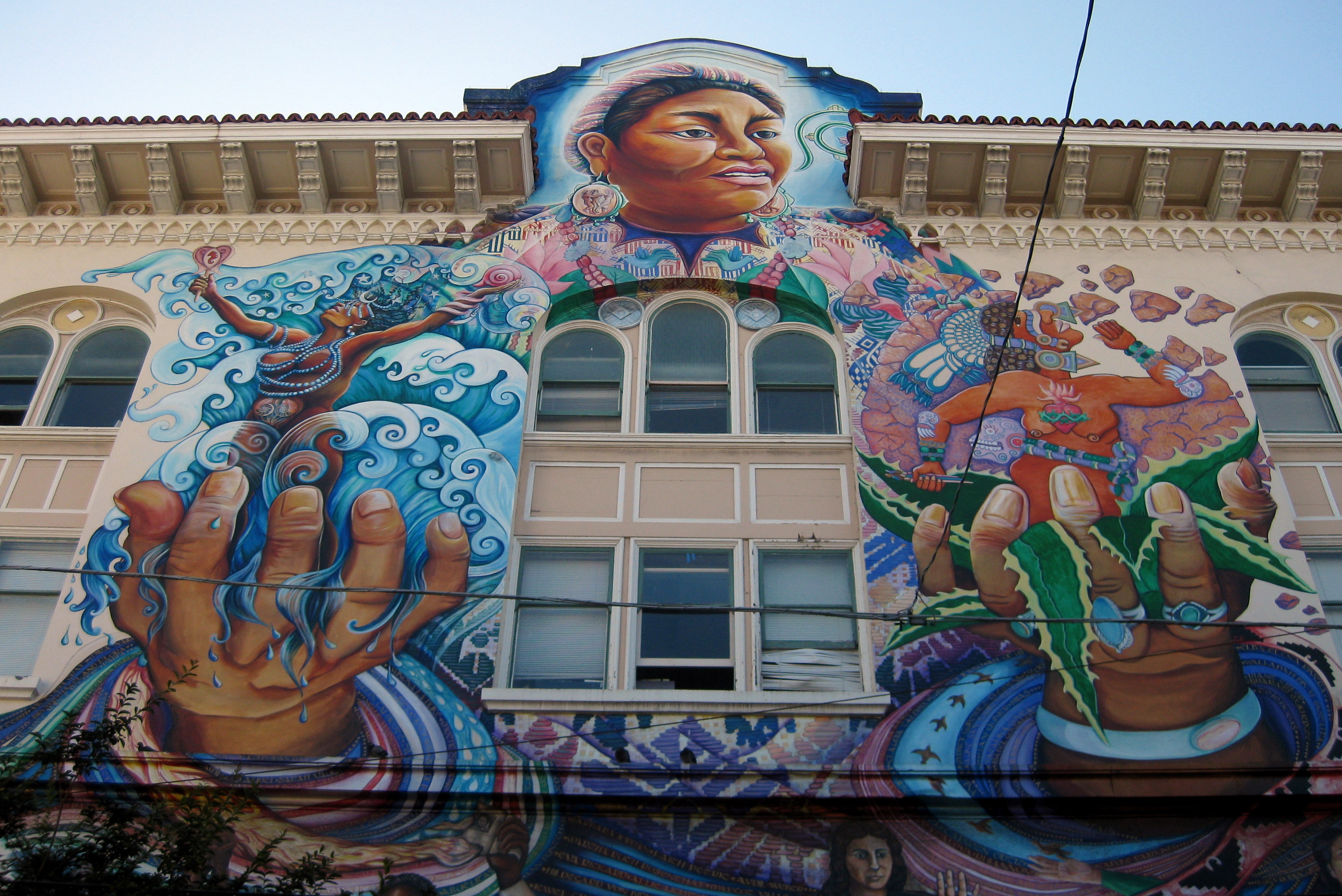 Colorful mural on a beige building with many windows featuring Rigoberta Menchu Tum holding Indigenous female deities Coyolxauhqui and Yemeyah, one in each of her oversized hands. 