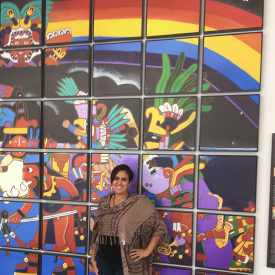Dr. Amber Rose González standing in front of a mural featuring a rainbow and Mesoamerican symbols