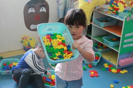 child holding a basket of multicolored toys up to the camera while another plays in the background