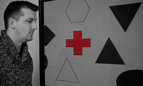 Une photographie montre une personne regardant fixement un écran qui affiche une croix rouge sur le côté gauche et de nombreuses formes en noir et blanc partout.