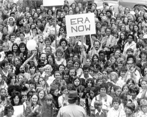 Black and white photo showing hundreds of women and men supporting the Equal Rights movement. Some are holding signs saying ERA NOW. Standing in front with their back turned to the camera is a police officer.