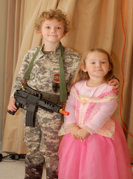 A little boy and girl are standing next to each other. The boy is dressed in a military uniform with a fake gun hanging around his neck. The girl is wearing a pink princess dress.