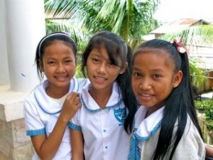 Three young Asian school girls, wearing white and blue school uniforms, smiling at the camera with their arms around each other.