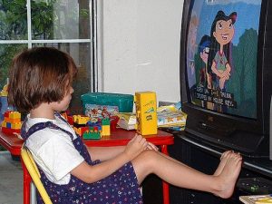 Preschool age girl sitting right in front of the TV with her legs propped up on the TV stand.
