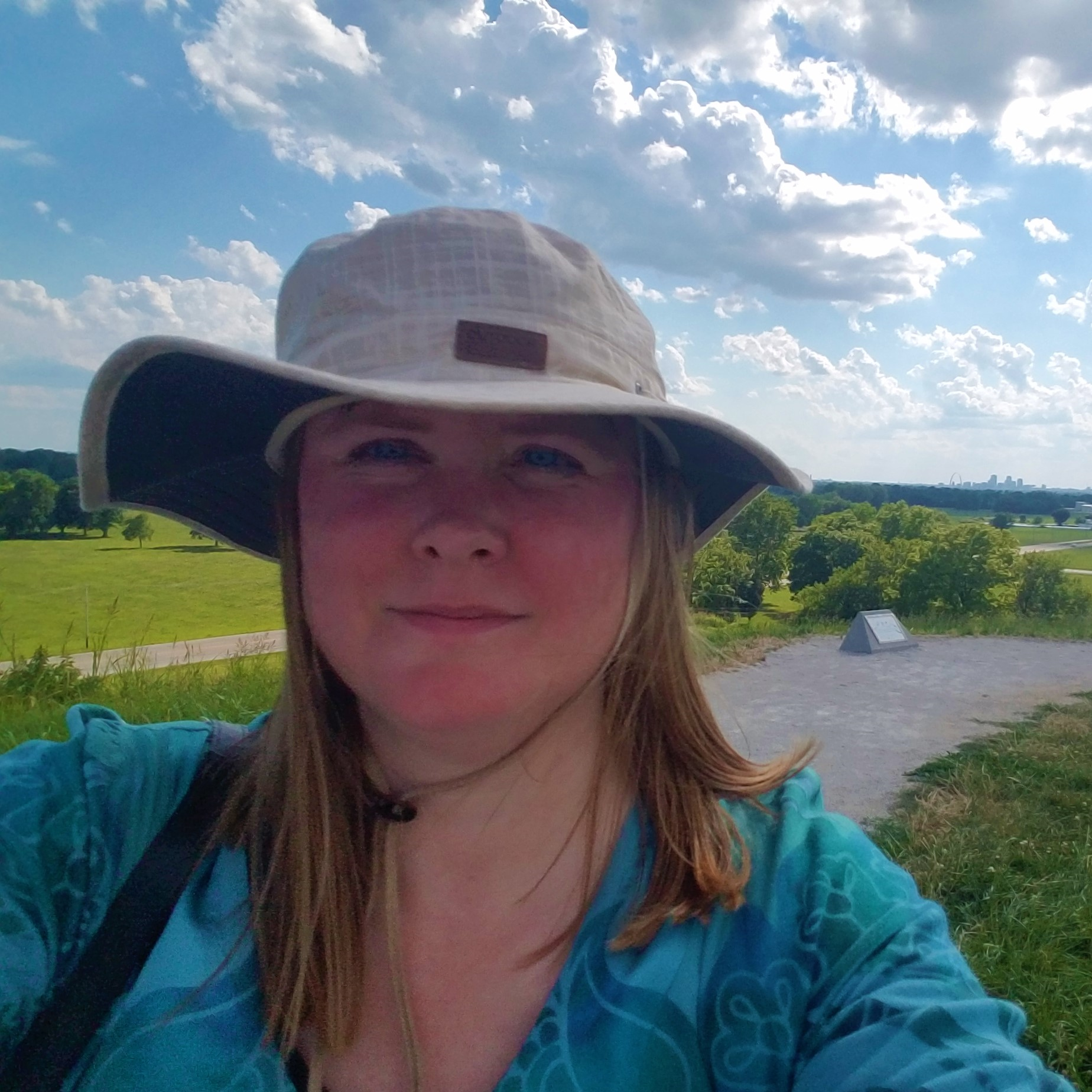 Selfie of blonde haired author wearing brimmed hat. An expanse of nature is the background.