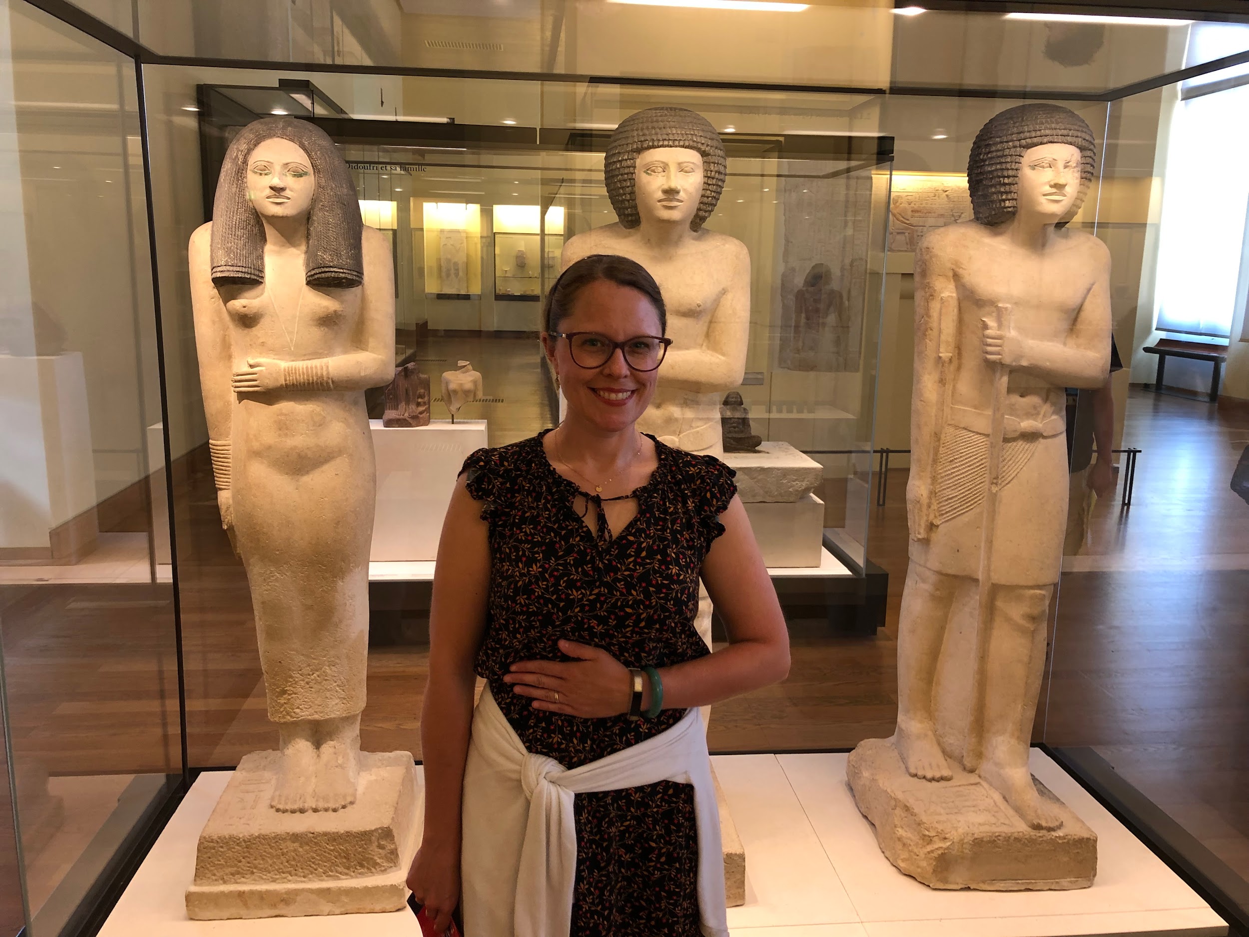 A woman stands in front of museum statues imitating the statues' gestures with a hand over the stomach.