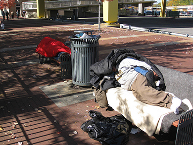 The image is a photograph of two people who are homeless and sleeping on public city benches.