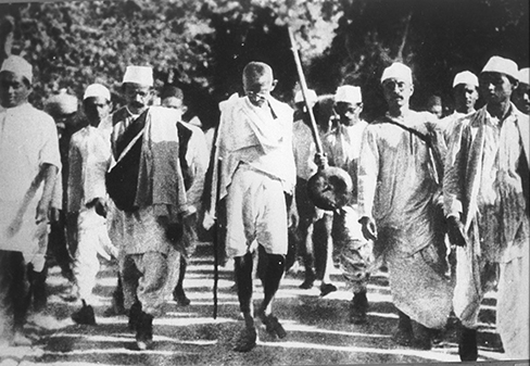Se muestra una fotografía de Mohandas Gandhi. Hay varias personas caminando con él.