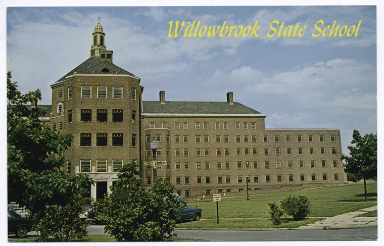 A postcard shows a brown brick institutional building with a road, cars, and street signs. There is a green lawn with trees and sidewalks and a blue sky with clouds. Written in yellow script are the words “Willowbrook State School.”