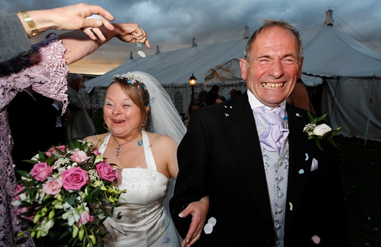 Portrait of a woman with Down's syndrome on her wedding day with her husband. Confetti is being thrown at the bride and groom who are smiling and laughing. 