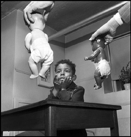 black and white image of a young Black child pointing at Black and white baby dolls being held by their heads in the hands on a white man; received in black frame