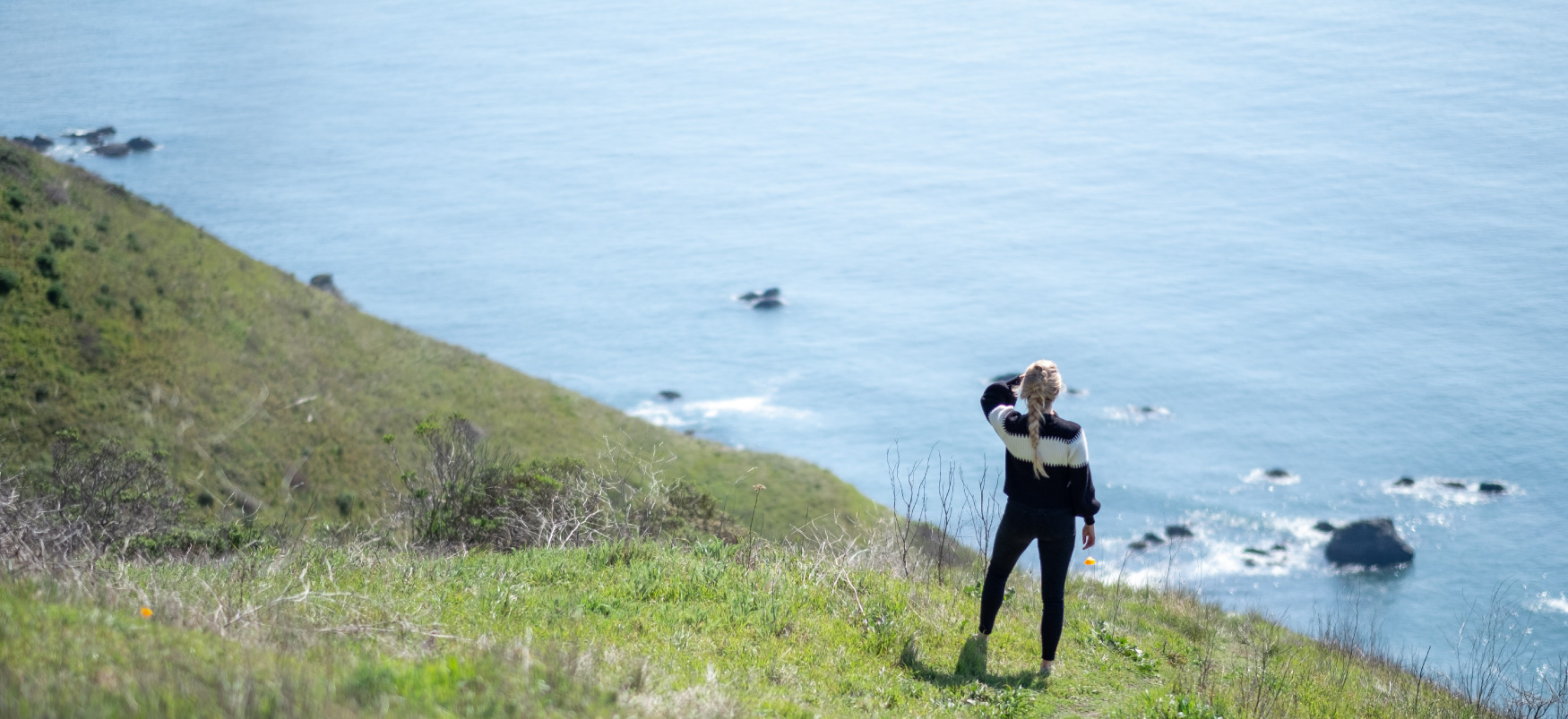 A person stands on a grassy hillside, shielding their eyes from the sun and looking out over the ocean.