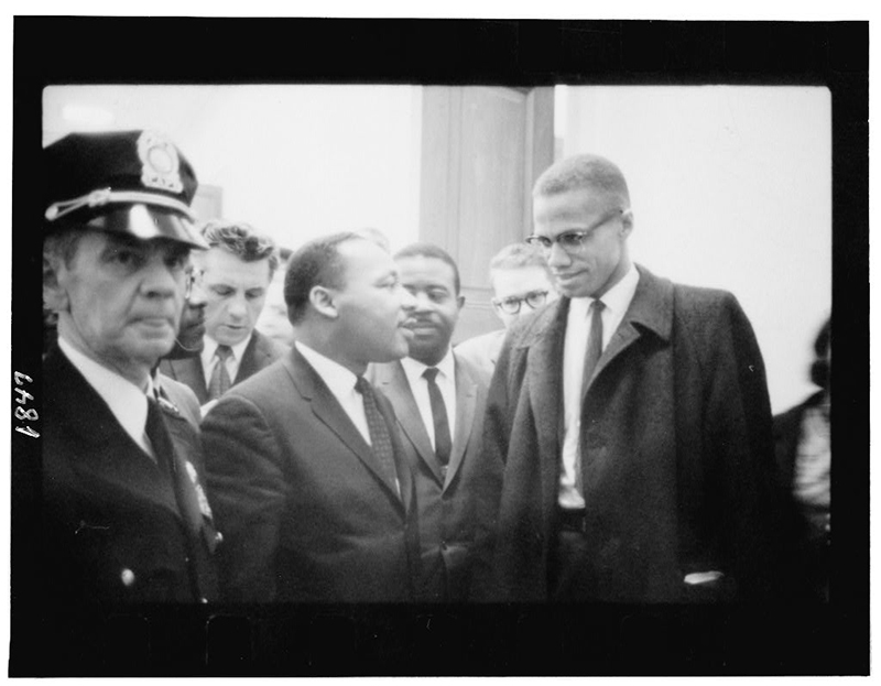 Martin Luther King Jr. stands next to Malcolm X. A uniformed police officer stands in the foreground next to Martin Luther King Jr.