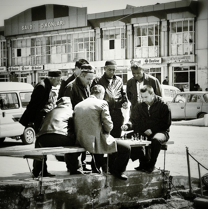 Two people play chess on a bench at the side of a street. A group of people surrounds the bench, watching the match.