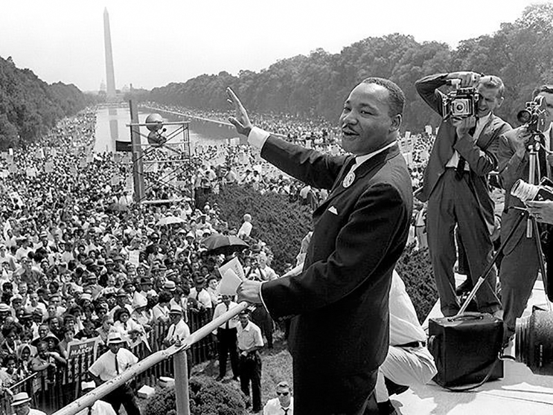 Rev. Martin Luther King Junior waving to a large crowd from a platform podium and being photographed by the press at the 1963 March on Washington.