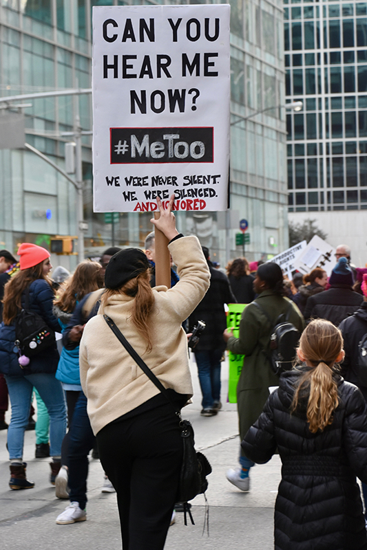 A woman walks among protesters on a city street carrying a signboard that reads “Can You Hear Me Now? #MeToo.”