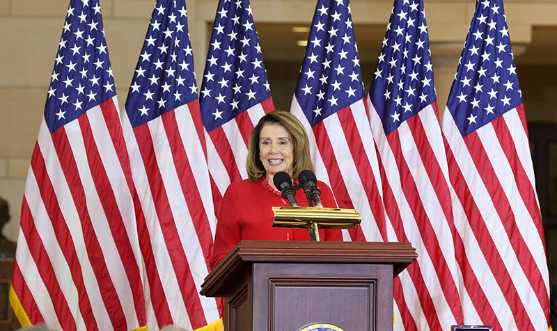 Nancy Pelosi stands before a podium, in front of six American flags, speaking into a microphone.