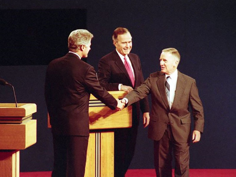 On a stage with two podiums, Bill Clinton shakes Ross Perot's hand while George H.W. Bush looks on.