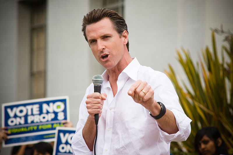California Governor Gavin Newsom speaks into a hand-held microphone as constituents hold up signs in the background that read “Vote no on Prop 8.”
