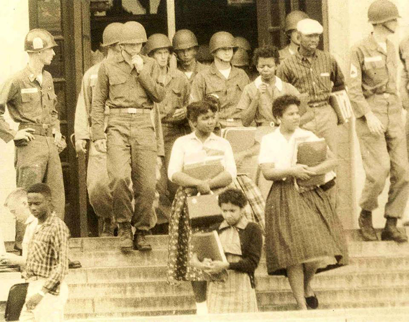 Black students walk down the stairs at the entrnace to a building, holding books in their arms, escorted by uniformed military personnel wearing combat helmets.