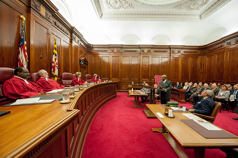 Five judges sit at the bench at the front of a court room. A man speaks from a podium in front of the judges. Additional people sit at tables on either side of the podium and in rows of chairs behind the tables.