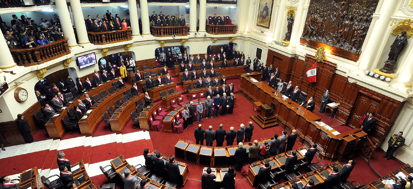 In a legislative chamber an assembly of professionally dressed people stand behind wooden desks, arranged in a tiered semicircle, facing a long rostrum, while additional people stand and look on from the gallery above.