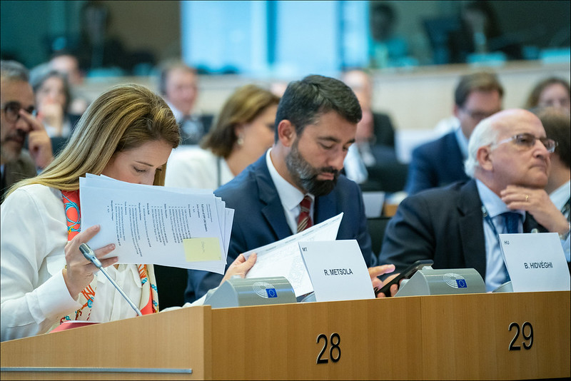 European Parliamentarians sit in numbered rows of connected wooden desks, facing forward, and in some cases looking through papers.