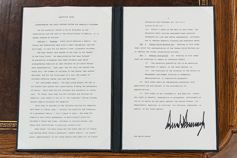 An open portfolio on a desk shows side-by-side sheets of paper typed with an executive order issued by the President of the United States and signed in the lower right by then-president Donald Trump.