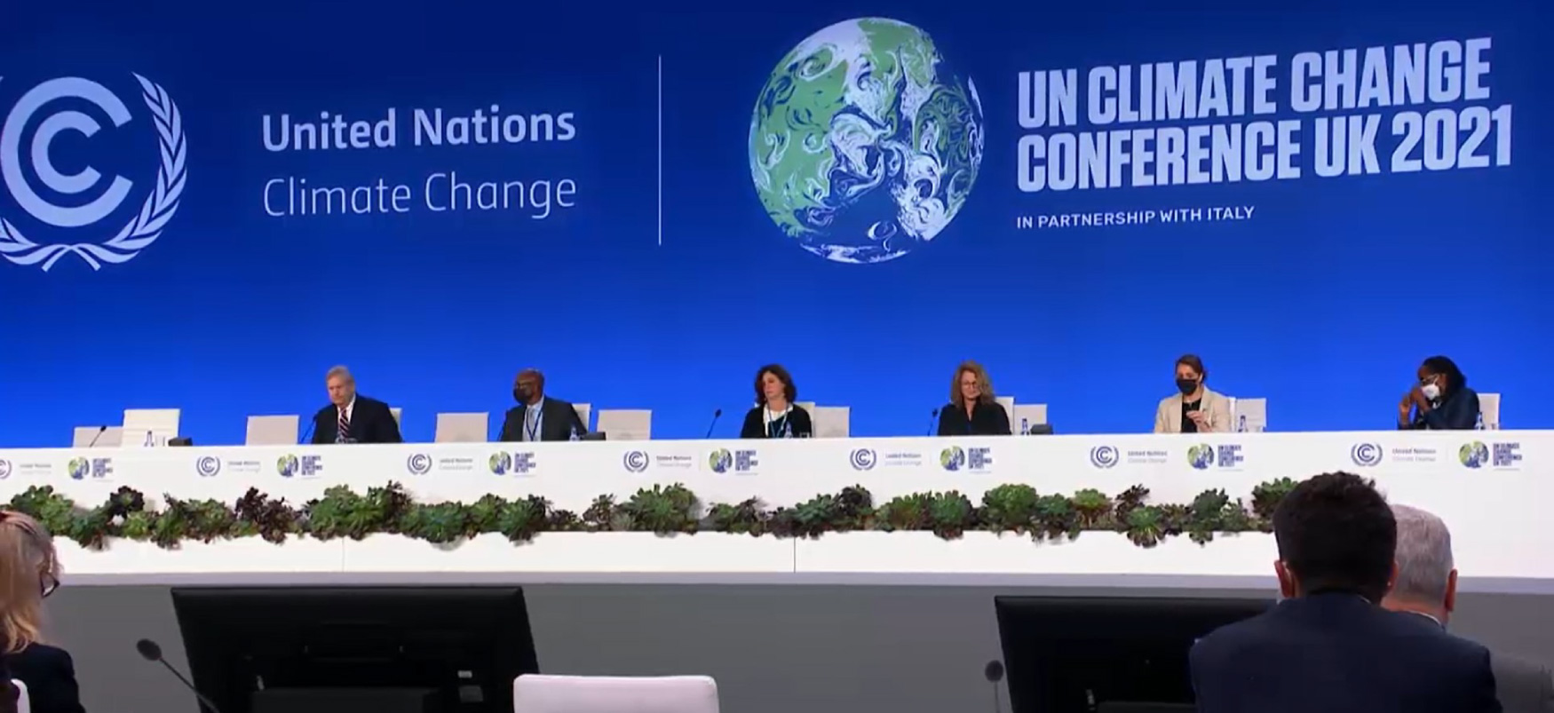 Six people sit on a dais behind a long white desk. On the wall behind them are the words “United Nations Climate Change” and “U N Climate Change Conference U K 2021 in partnership with Italy.” A drawing of the Earth is also on the wall.