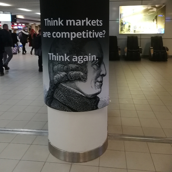 An advertisement shows the words “Think markets are competitive? Think again” across a black and white portrait of Adam Smith.