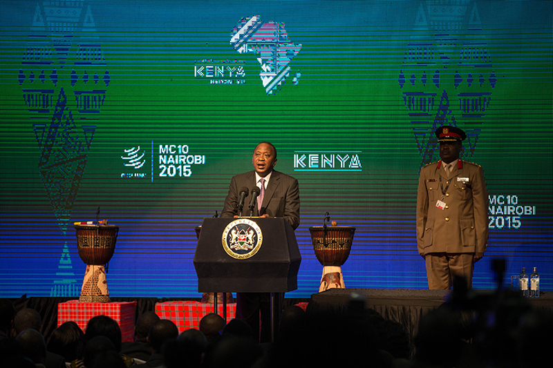 A person stands at a podium, giving a speech. Behind this person, another person in a military uniform stands at attention. The wall in the background reads “M C 10 Nairobi 2015” and “Kenya.”