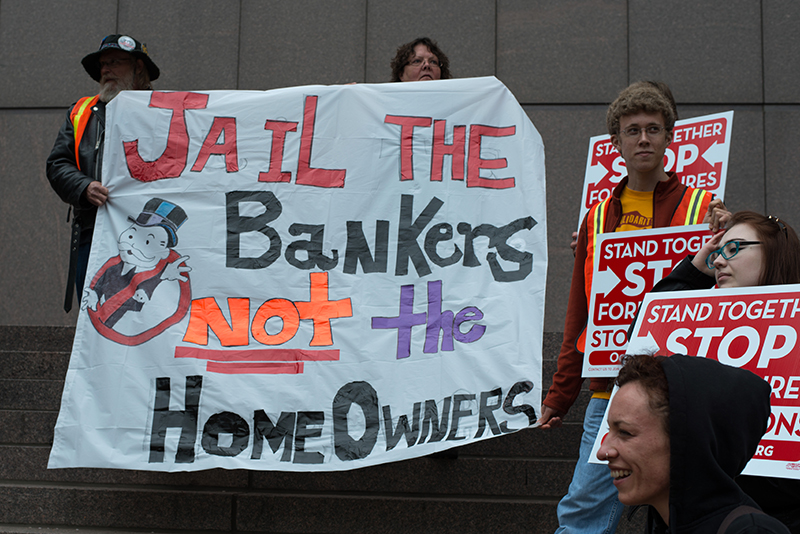 Five protestors hold up signs. The largest sign says “Jail the bankers not the homeowners.”