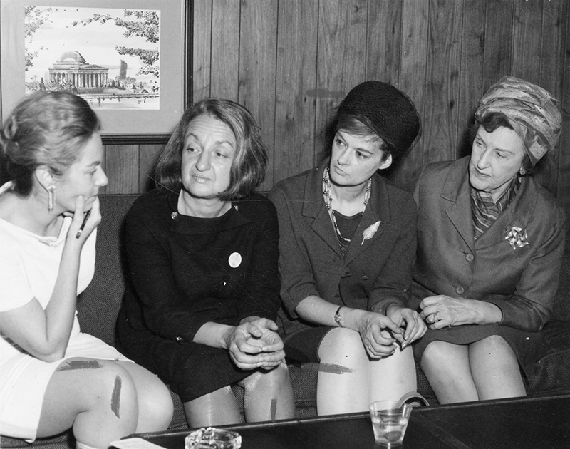 Four women sit on a couch talking. Betty Friedan is the second from the left. The three women on the right lean in to listen to what the woman on the left is saying.