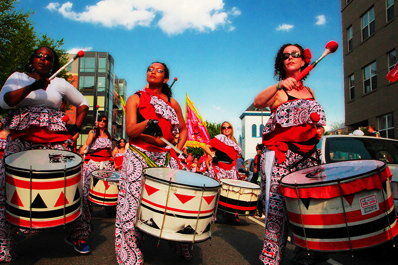 A group of woman wearing red, black, and white patterned clothes play large drums in the street.