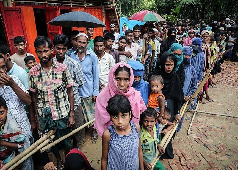A large group of Rohingya men, women, and children stand in rows outside of a wooden building.
