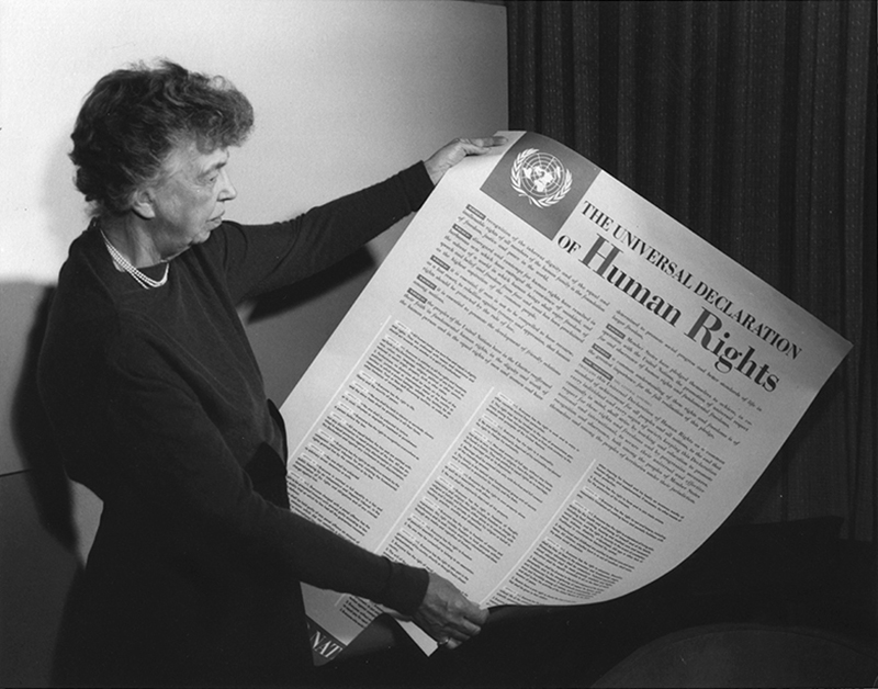 Eleanor Roosevelt displays a large poster with the text of the Universal Declaration of Human Rights in English.