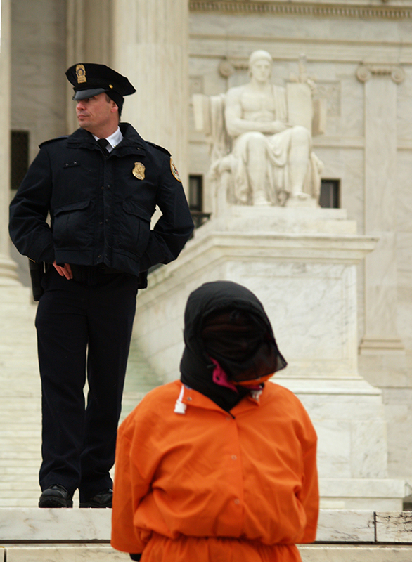 A person wearing an orange jumpsuit stands in front of a marble building that is decorated with a statue of a seated figure and Greek columns. A black hood completely covers the person's head and face. Behind the person, a uniformed police officer stands on the steps of the building.