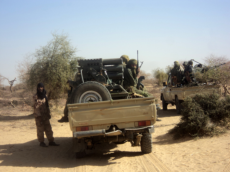 Two groups of people dressed in combat fatigues and cloth head coverings sit in the back of two pickup trucks in a dry, sandy landscape. Several of the people hold long guns. Another person stands to the left, wearing light brown camouflage and a dark cloth head covering.