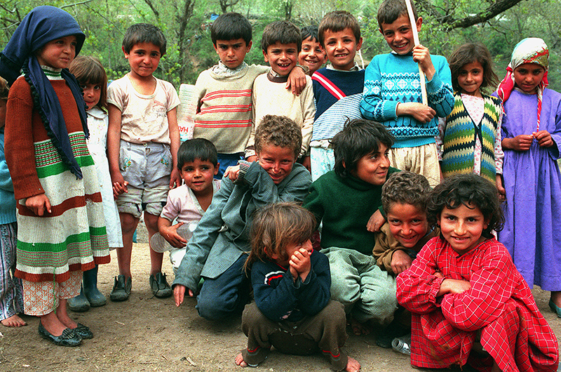 A group of children gathers to have their picture taken in an open area outdoors in front of dense trees. Some children crouch on the ground and some stand behind them.