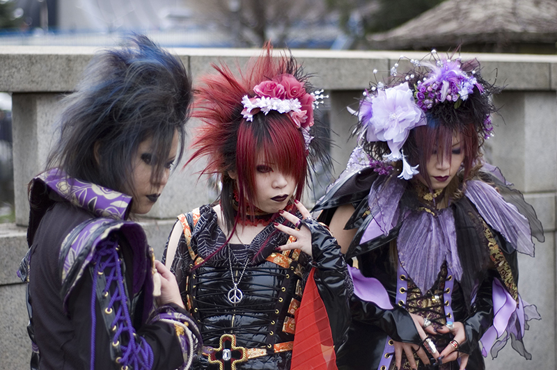 A group of three Japanese young people wear dark tailored or corseted clothes in shades of black, blue, red, and purple. They wear floral accents in their hair, which is teased out and dyed or streaked with colors that coordinate with their clothes. They also wear dark, dramatic eye and lip makeup.