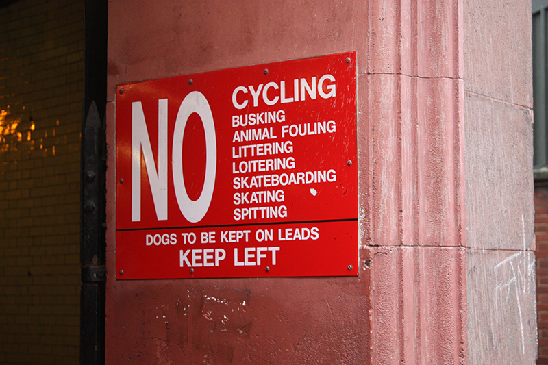 A red sign on a wall reads, “No cycling, busking, animal fouling, littering, loitering, skateboarding, skating, spitting.” It also reads, “Dogs to be kept on leads; keep left.”