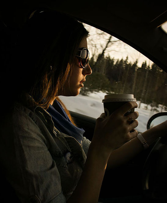 A person wearing sunglasses sits in the driver's seat of a car with one hand on the steering wheel while the other holds a to-go coffee cup.