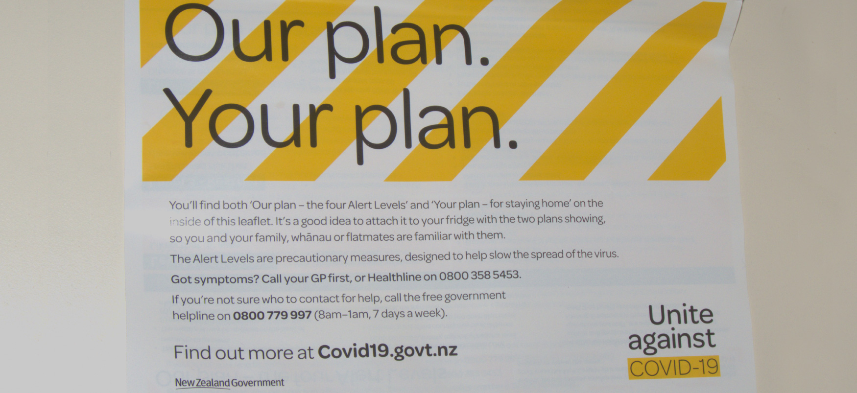 A poster hung on a wall reads “Our plan. Your plan.” against a backdrop of yellow diagonal stripes. The poster conveys information on alert levels and guidance on social distancing during the COVID-19 pandemic. It indicates that more information is available at Covid19.govt.nz.