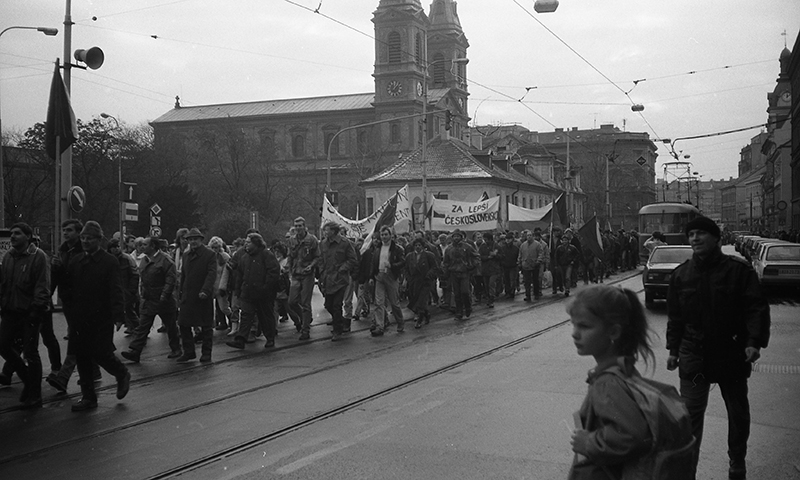 A black and white photograph shows people marching down the middle of a street, along street car tracks, carrying cloth banners.