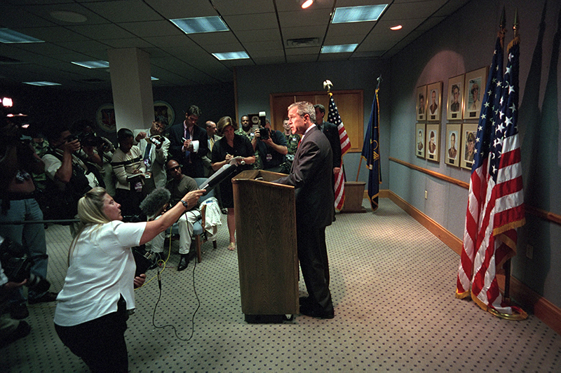 President George W. Bush speaks at a lectern in a room full of reporters. Three large American flags stand behind him.