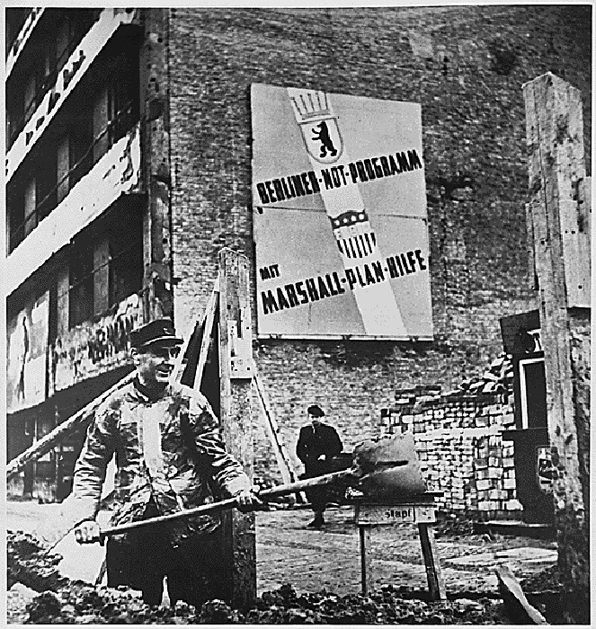 A black and white photograph shows a man shoveling rubble outside a damaged brick building. In the center of the outside wall of the building hangs a large sign written in German that includes the words “Marshall Plan.”