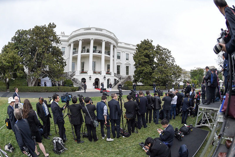 A large group of reporters wait in a long line outside the White House, many of them holding audio and visual recording equipment.