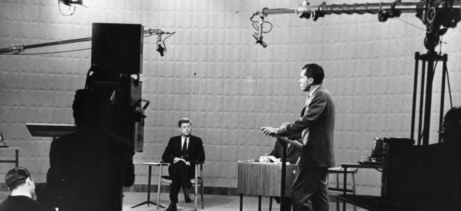 Republican Vice President Richard Nixon stands at a podium on a soundstage, facing a television camera, with microphones visible above his head. His seated opponent, Democratic Senator John F. Kennedy, looks on.