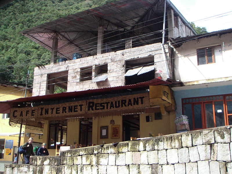 A multi-level structure with rough openings for windows and a metal roof sits along the side of a treed mountain road. A sign on the front of the structure says CAFE INTERNET RESTAURANT. Two backpackers walk up the road in the foreground.